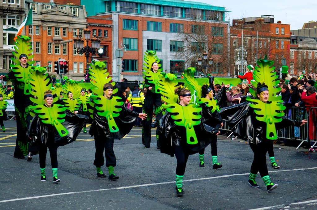 Dancers in green leaf costumes march down a Dublin street past a crowd, with an Irish flag above