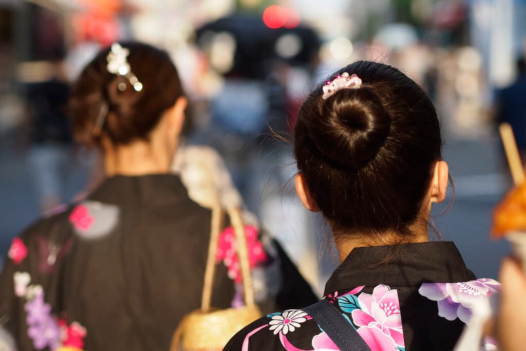 Two women in floral yukata with their hair pinned up, seen from behind, walk through a crowd on the evening of the Sumida River fireworks