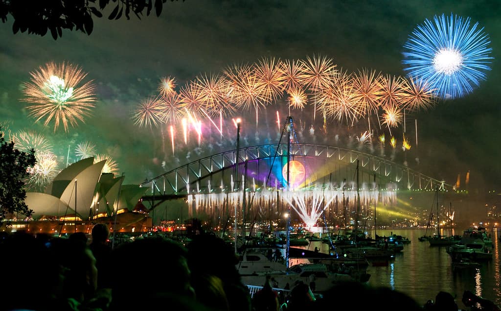 Fireworks burst over the Sydney Opera House and Harbour Bridge at night, with boats and a crowd in the foreground
