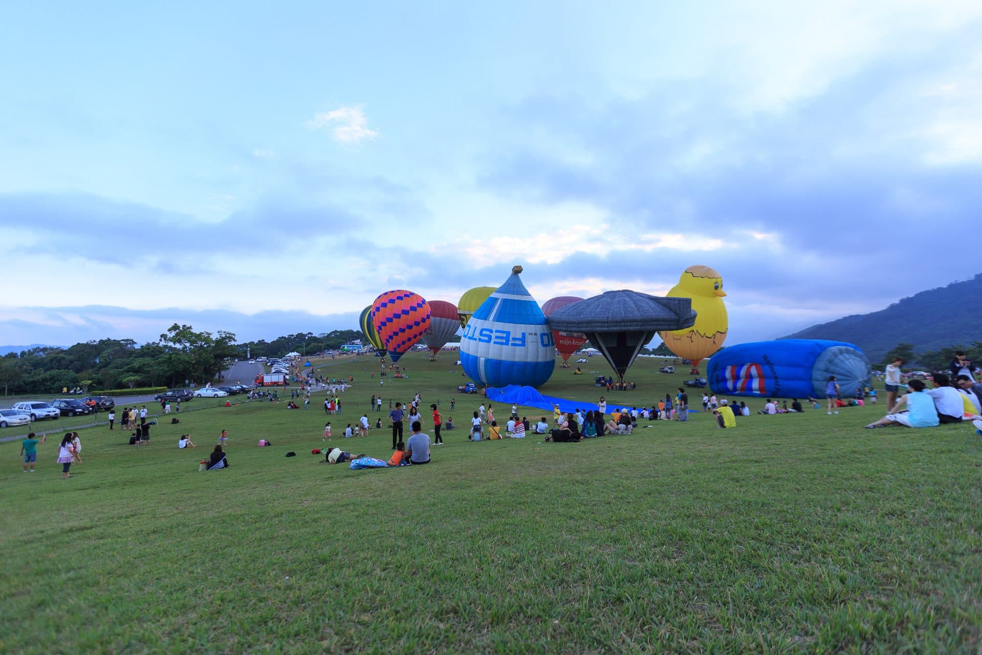 Hot air balloons, including a yellow chick and a blue-and-white drop shape, stand inflated on a grassy hillside as families watch