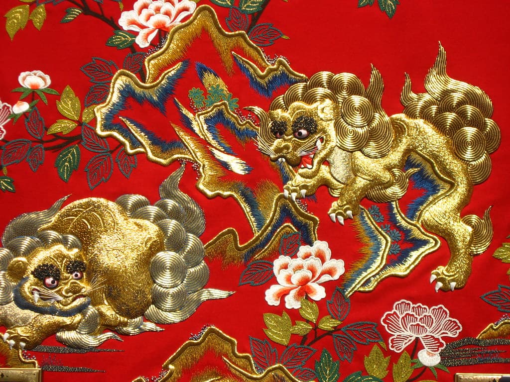 Gold-thread embroidery of two lion-dogs among peonies on a red festival cart panel