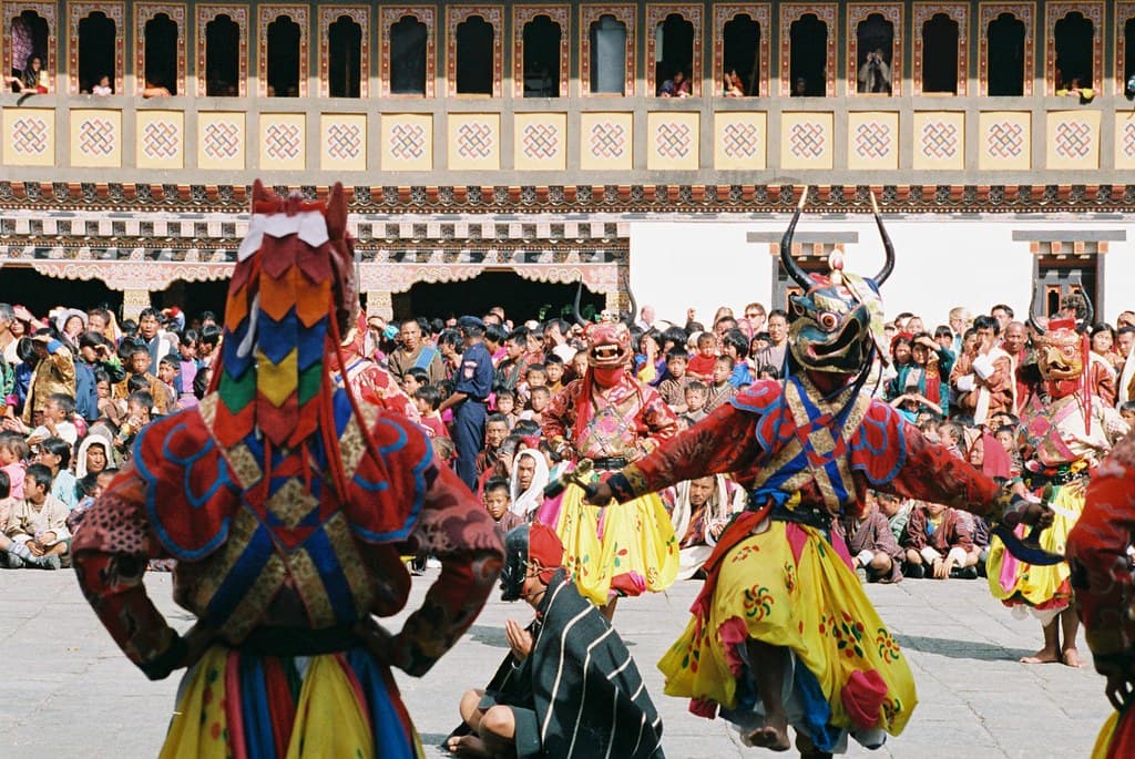 Mask dancers in bright silk costumes and carved masks perform in the courtyard of Tashichho Dzong, with crowds behind