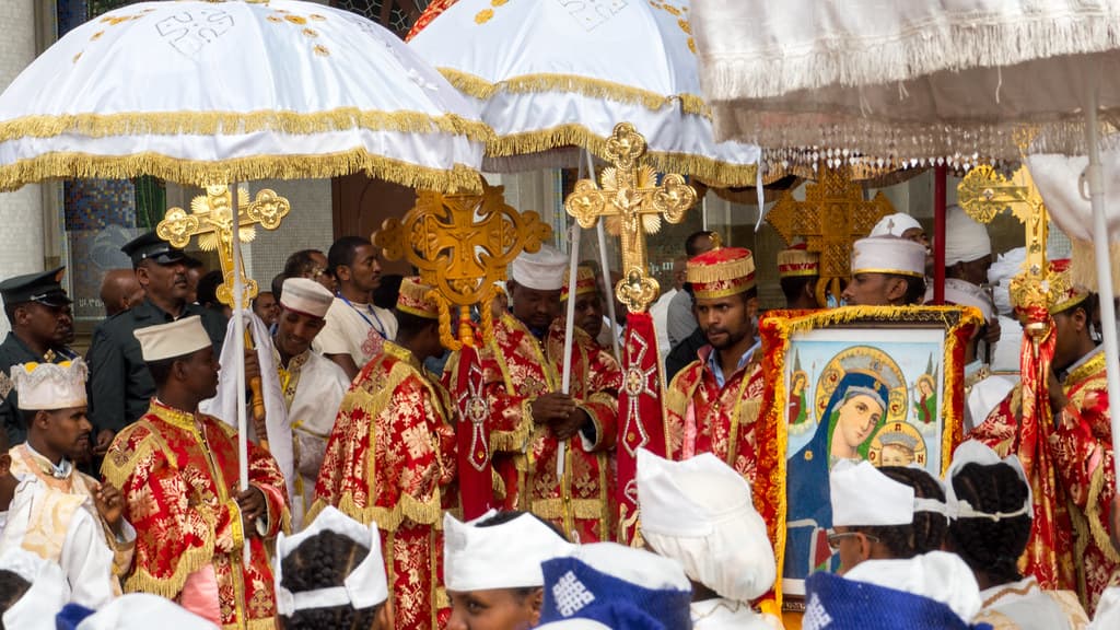 Clergy in red and gold robes carry processional crosses and a framed icon under white and gold umbrellas