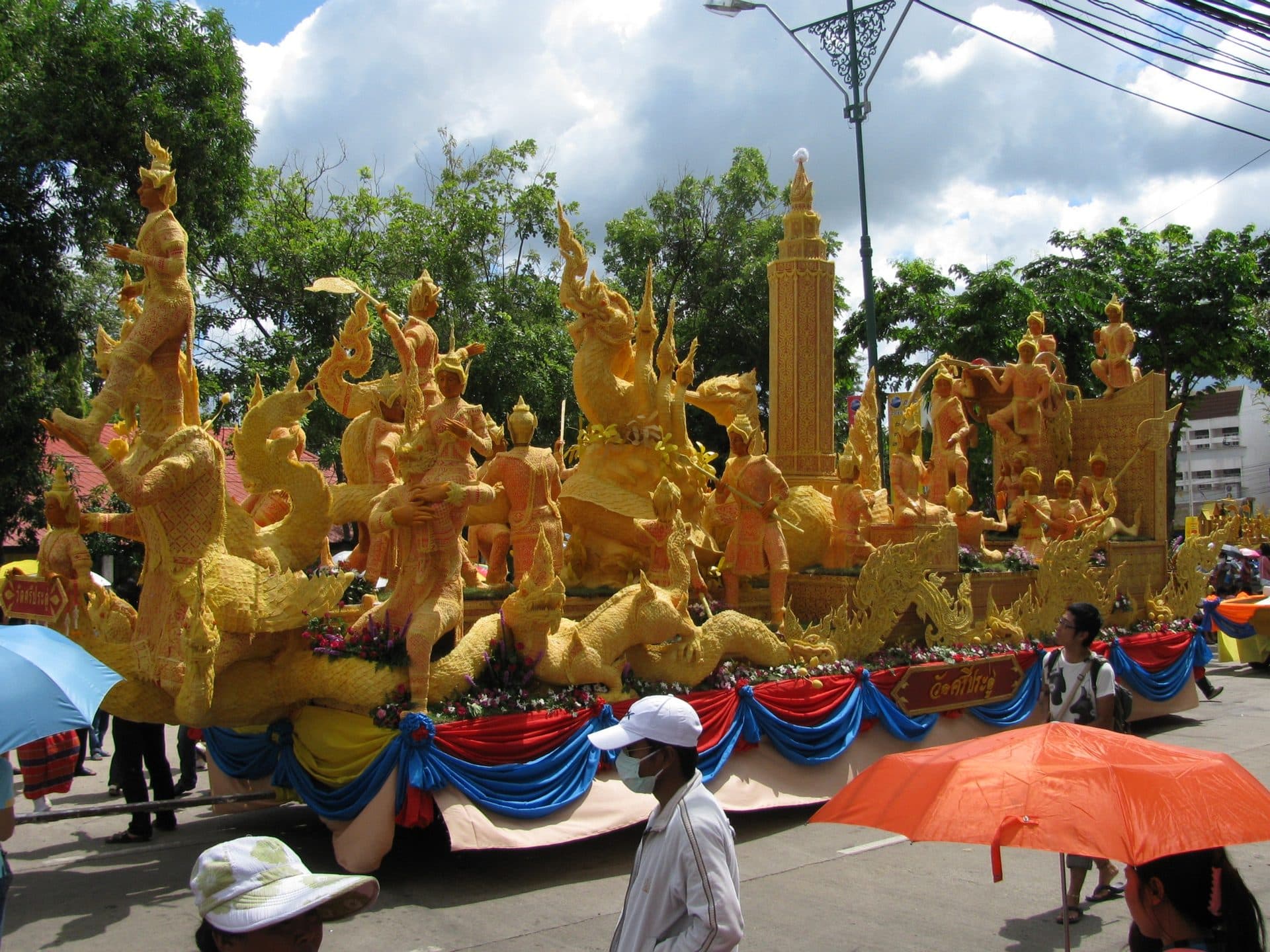 A tall wax-carved float of gilded figures and naga serpents on a decorated street float, with spectators passing in front