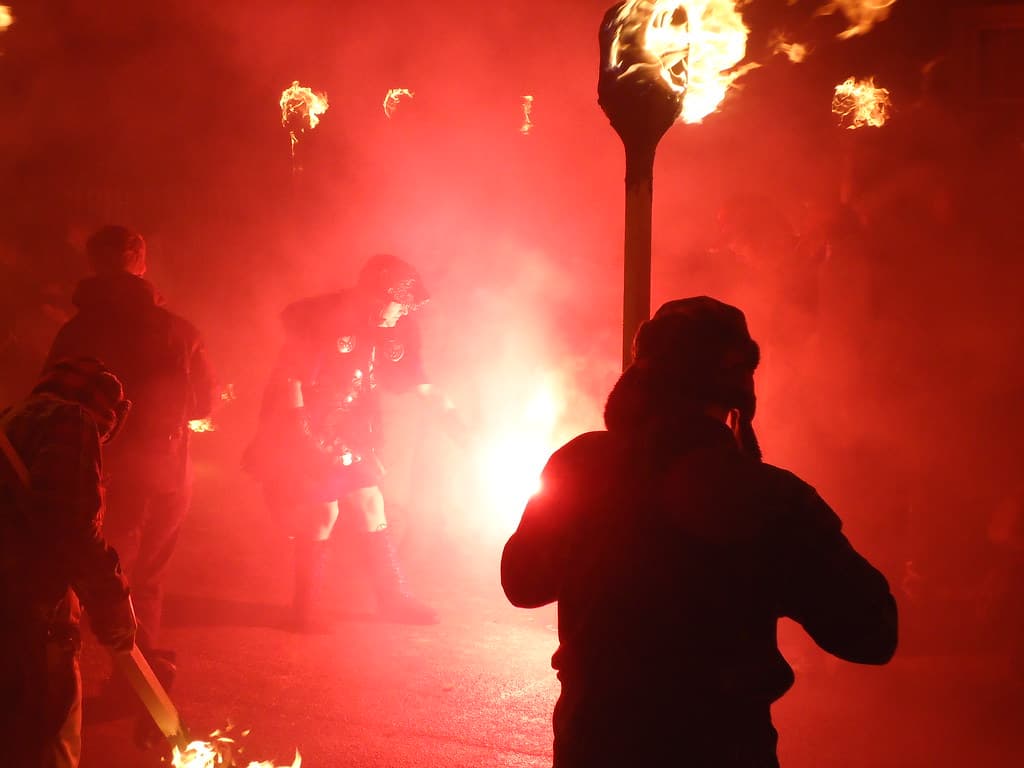 Costumed guizers and torch-bearers in red smoke at night, with flames on torches overhead