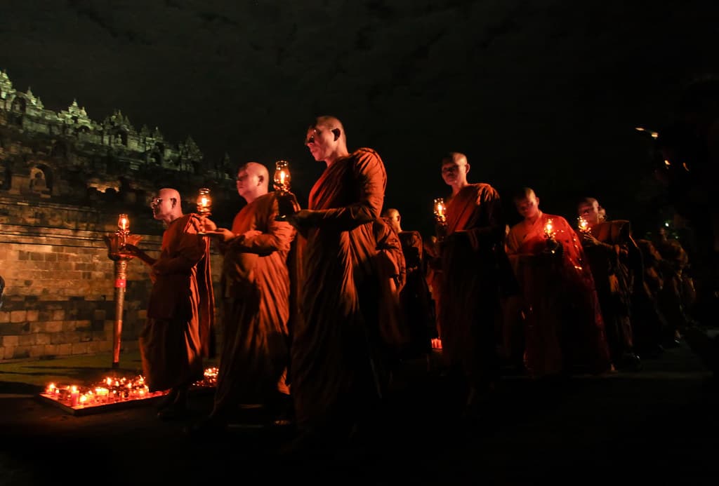 Buddhist monks in saffron robes carry oil lamps in a night procession beside the stone walls of Borobudur