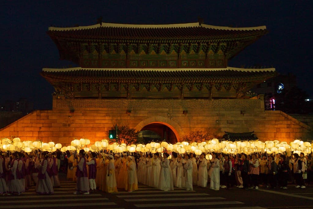 People in hanbok carry glowing lanterns in a night procession in front of a historic gate
