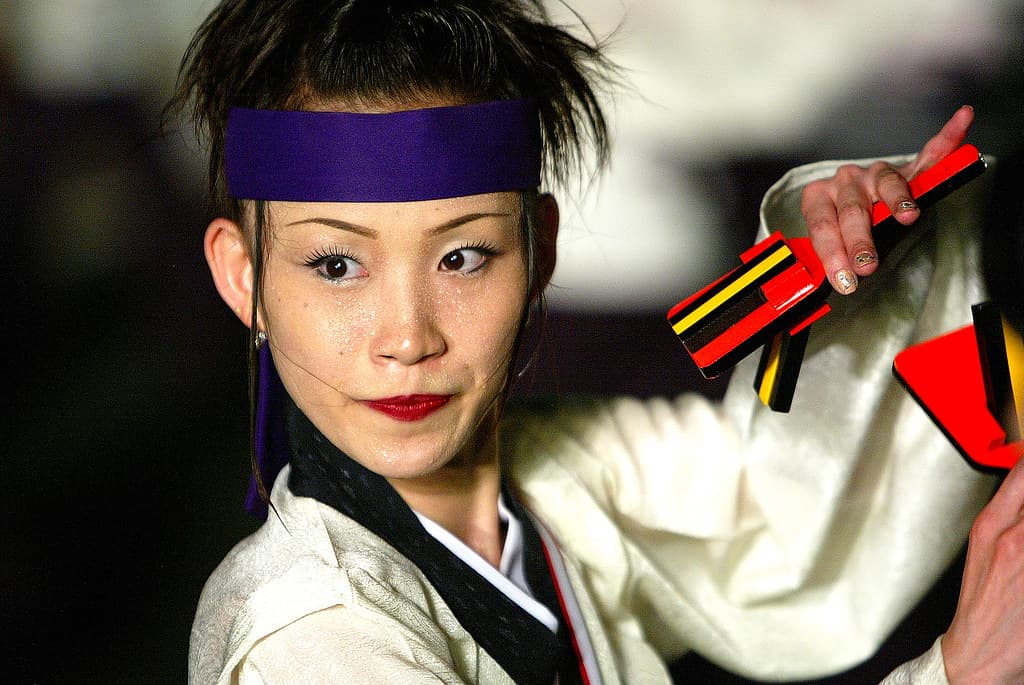 A yosakoi dancer with a purple headband and white costume holds red and yellow naruko clappers at the Kochi festival