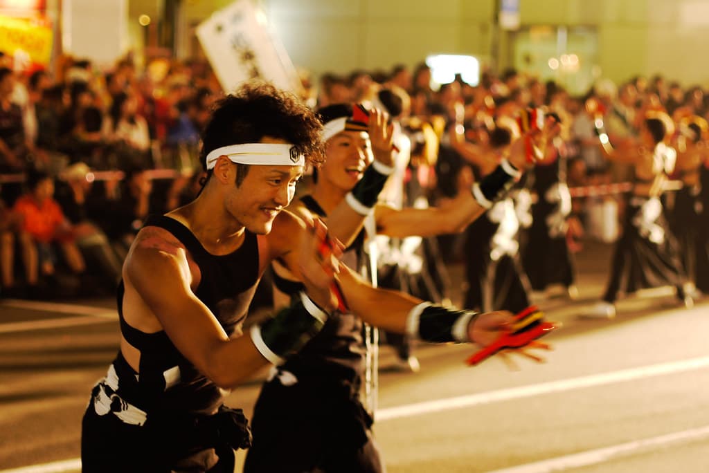 Dancers in black costumes with red armbands perform in formation along a street in Sapporo, watched by crowds